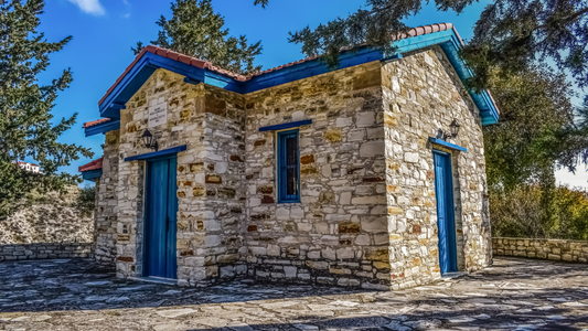 A small, well-preserved historic stone building with blue doors and trim, showcasing traditional masonry techniques under bright daylight in a tree-lined setting.