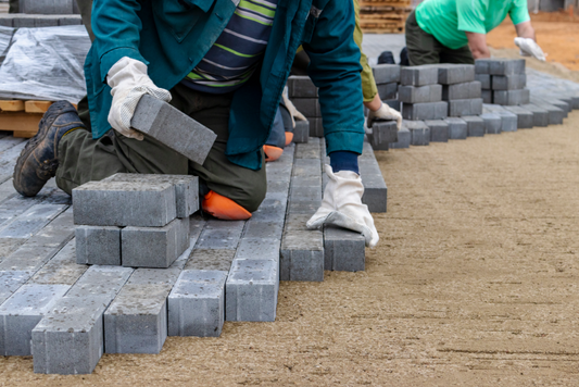 Construction workers carefully laying gray paver bricks on a prepared sand base for a patio or walkway, showcasing skilled masonry techniques and attention to detail.